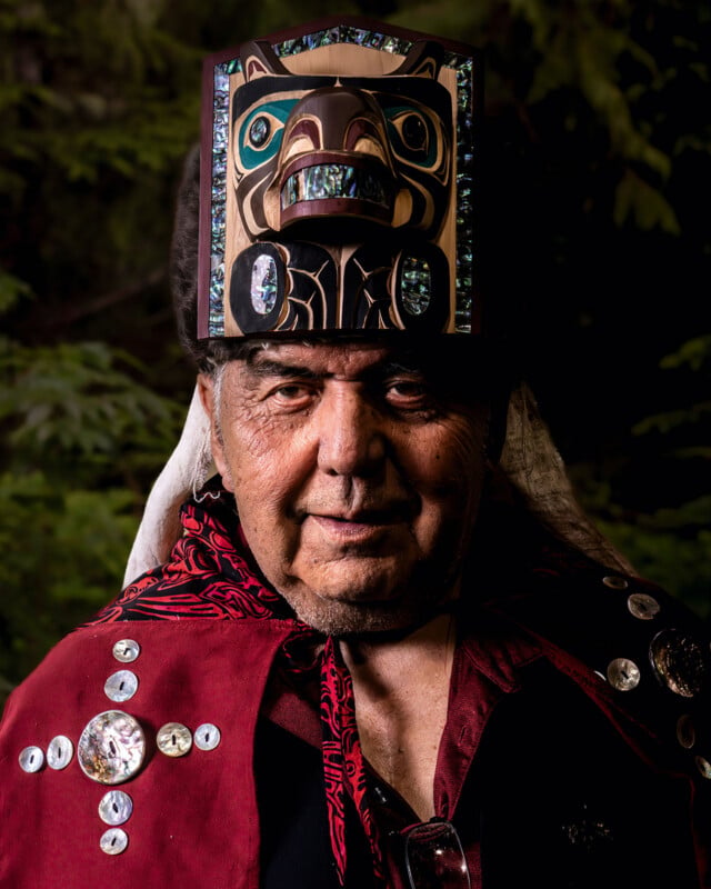 An elder wearing a traditional robe decorated with decorative buttons and a carved and painted wooden headdress stands in front of a background of blurred leaves.