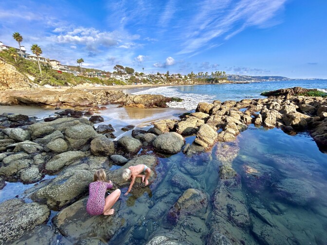 Two young white-skinned children play in a tidal pool under a blue sky on a sunny day. 