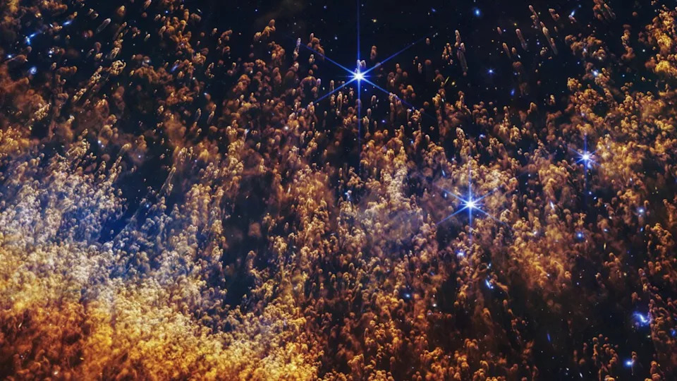 Hundreds of golden and orange clouds trail feather-like trails behind them. Small clouds cover a few scattered bright stars. 