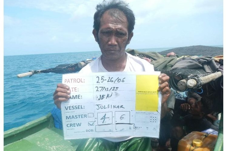 A man in a white shirt is sitting facing the camera and holding a sign with his personal information written on it.
