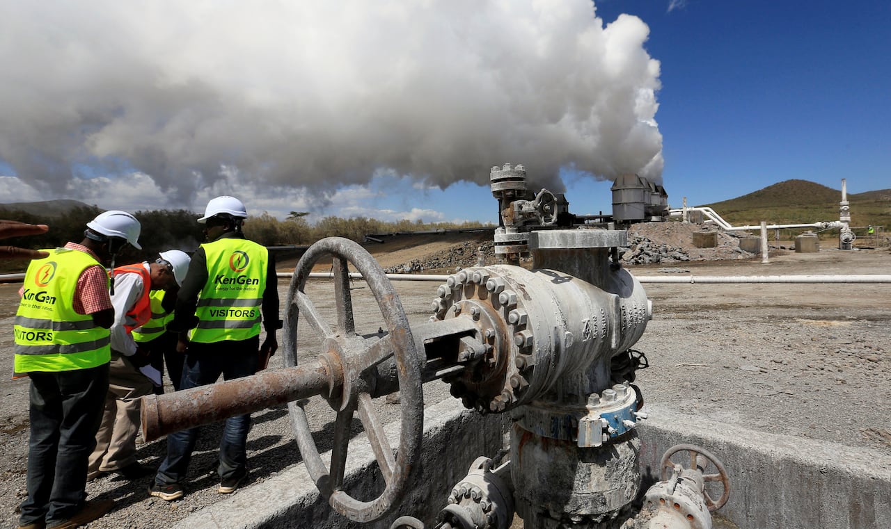 Men in high-visibility vests and helmets stand near metal wheels and pipes as steam billows from distant cylinders.