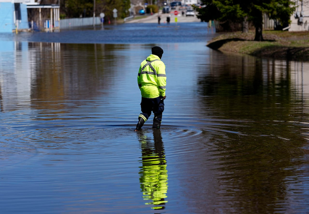 A worker in a bright yellow jacket is walking through a flooded city street on a sunny day.