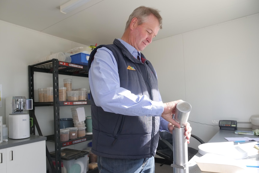 A man with gray hair, a demin shirt, and a dark blue vest pours lentils into a silver container on a minilab bench.