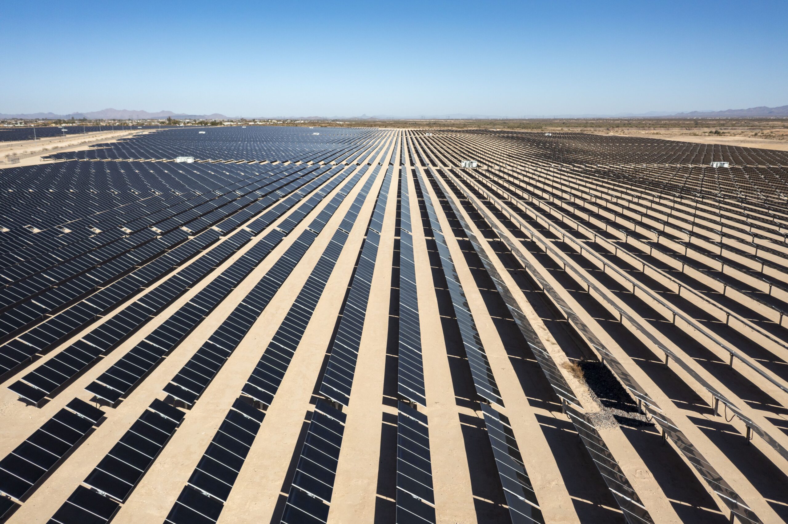 A vast solar panel farm is located in an arid desert region under clear blue skies and mountains on the horizon.