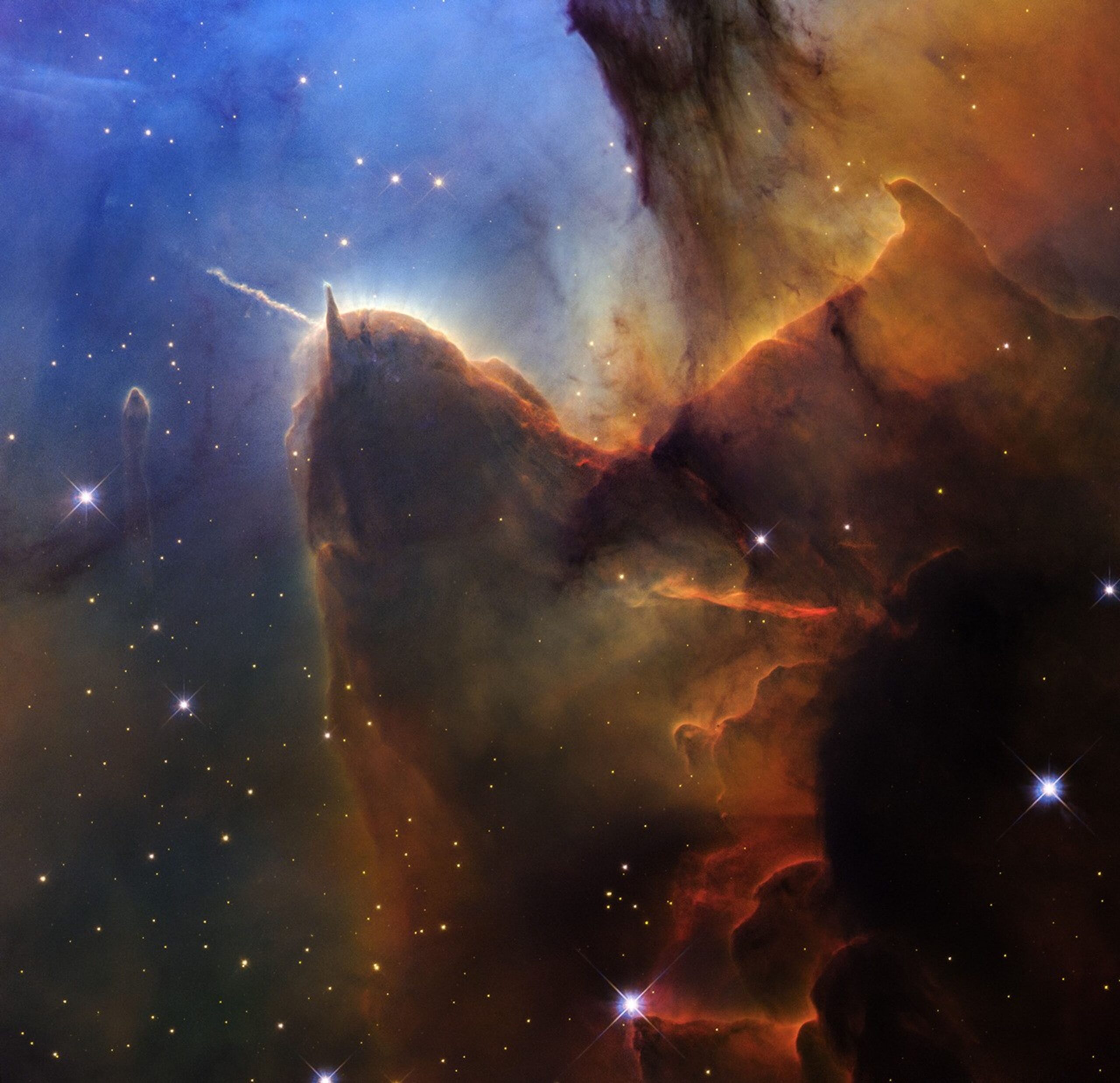 Hubble's view of the vast star-forming region known as the Trifid Nebula. The upper left is bright blue. Brown and amber draw irregularly overlapping lines from the top right through the center and flow down to the bottom center. The bottom right corner is almost completely dark. Small amber stars appear throughout the scene. On the left side, a brown shape stands out that looks like a head with two horns. The left corner points to the left and is wavy. The right corner is triangular and points upwards. The brown dust flows down the back and continues toward the upper right. A pronounced line appears below the center of the body, approximately the same length as the left horn, changing from orange to red. On the left side of the head there is a separate small translucent column. You can see some slightly larger blue foreground stars with four diffraction spikes.