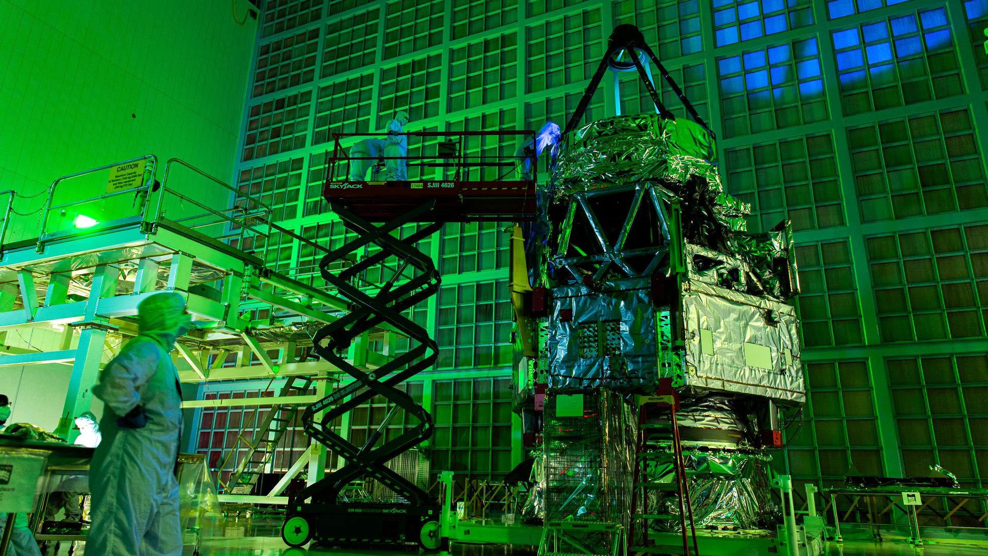 A telescope with a triangular top wrapped in foil stands next to scaffolding, and people in clean white suits observe it under green light.