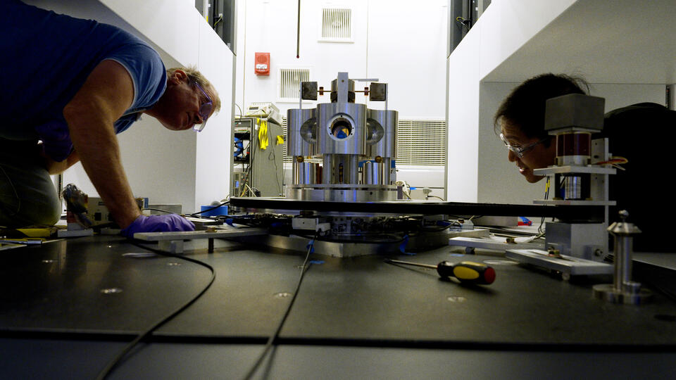 A wide shot shows two researchers on either side of a scientific device made of metal cylinders, staring intently at it.