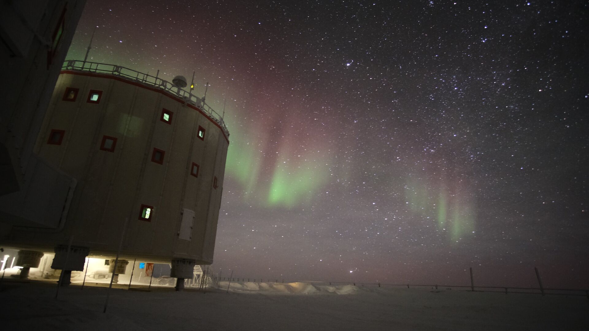 A cylindrical building with many small windows. Southern light dances in the sky behind you, creating ribbons of green and magenta against the starry sky.