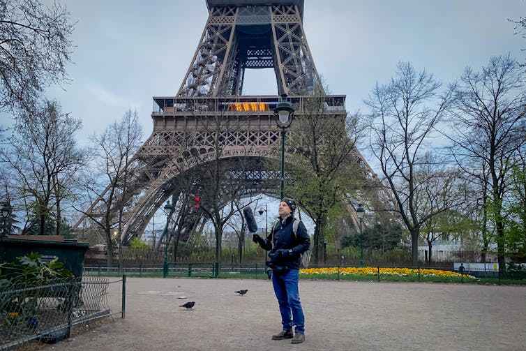 The researcher stands with the Eiffel Tower behind him, holding out a large black microphone.